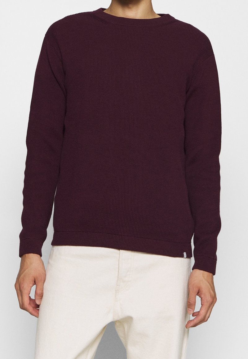 Homme portant un pull à manches longues côtelé violet foncé et un pantalon écru, debout devant un fond clair uni.