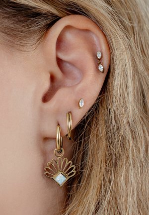 Nahaufnahme eines Ohrs mit hellbraunem Haar, das goldene Creolen und kleine goldene Ohrstecker in Diamantform trägt.