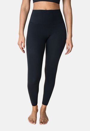 Woman wearing high-waist black leggings and a matching crop top, standing barefoot against a plain background.