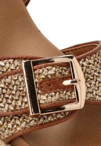 Sandal con tira trenzada marrón que presenta una hebilla de metal dorado, tela texturizada y una plantilla suave, ideal para el uso casual.