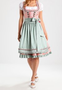 Femme portant une robe dirndl traditionnelle rose avec blouse blanche, tablier vert noué en nœud, et ballerines blanches.