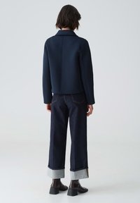 Giacca su misura blu navy con colletto e maniche lunghe, abbinata a jeans in denim scuro con orli risvoltati e una vestibilità rilassata.