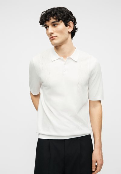 Νέος άνδρας με σγουρά σκούρα μαλλιά, φορώντας λευκό πόλο με κοντά μανίκια και μαύρο παντελόνι, κοιτάζοντας ελαφρώς προς το πλάι με τα χέρια πίσω από την πλάτη.