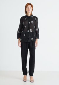 Bluză neagră transparentă cu nasturi, decorată cu modele abstracte albe, purtată cu pantaloni negri largi. Modelul poartă sandale cu toc transparent.