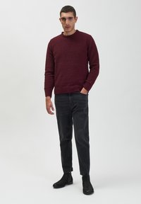 Burgundowy sweter dzianinowy z okrągłym dekoltem, noszony z czarnymi jeansami slim-fit i czarnymi botkami za kostkę. Teksturowana tkanina, prążkowane mankiety i dół.
