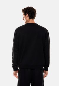 Černá mikina s dlouh�ými rukávy, žebrovanými manžetami a lemem. Má kulatý výstřih a loga na rukávech. Hladká textura.