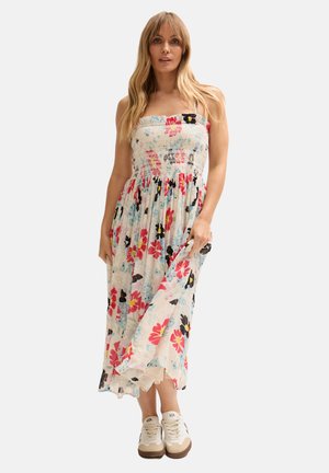 Frau mit langen blonden Haaren, die ein ärmelloses weißes Midikleid mit Blumenmuster und beige Sneakers trägt, hält den Saum des Kleides.