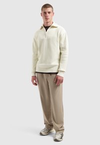 Junger Mann steht, trägt einen cremefarbenen Half-Zip-Pullover, beigen Hosen und silbernen Laufschuhen vor einem schlichten hellgrauen Hintergrund.