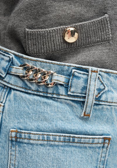 Κοντινό πλάνο ανοιχτό μπλε τζιν με ασημένια ζώνη αλυσίδας και μέρος από γκρι πλεκτό πουλόβερ με ένα χρυσό κουμπί.