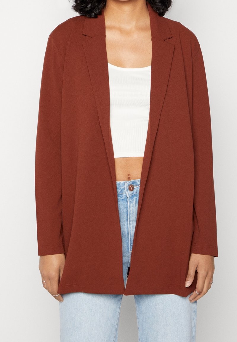 Hnědý blazer s volným střihem, dlouhými rukávy a klopami, nošený přes bílý crop top, doplněný o světle modré džíny.