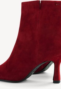 Bottine en daim rouge avec fermeture éclair latérale et talon stiletto incurvé de hauteur moyenne sur fond blanc.