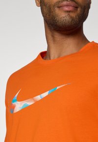 Camiseta de algodón naranja con un logo de Nike en blanco y azul con patrón en el pecho. Cuello redondeado y mangas cortas.