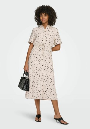 Nő bézs színű, rövid ujjú, gombos midi ruhát visel fekete pöttyökkel, fekete kézitáskát tart, és fekete magassarkú szandált visel.