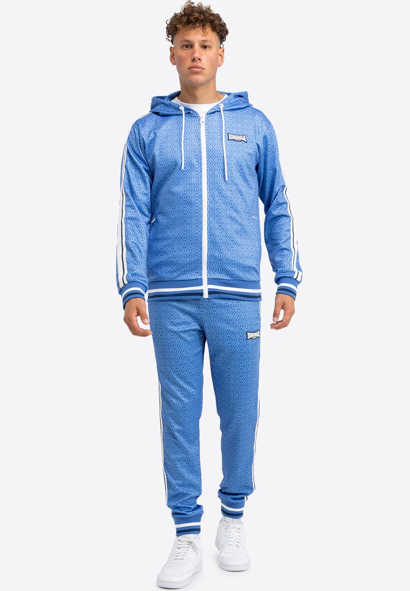 Blauer Sportanzug mit geometrischem Muster, kontrastierenden weißen Streifen, Kapuzenjacke mit Reißverschluss und elastischen Bündchen. Kombiniert mit weißen Sneakern.