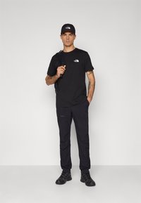 Tricou negru și pantaloni dintr-un material ușor, cu logo-ul mărcii. Accesoriile includ o șapcă neagră și un rucsac. Pantofii negri completează ținuta.