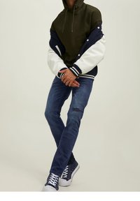 Huvtröja i olivgrön med ärmar i marinblått och vitt i varsity-stil, kombinerad med slitna blå jeans och svarta sneakers.