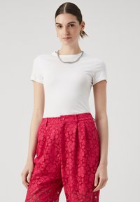 T-shirt bianco a maniche corte abbinato a pantaloni in pizzo rosa brillante con motivi floreali, dettaglio a bottoni in vita e finitura testurizzata.