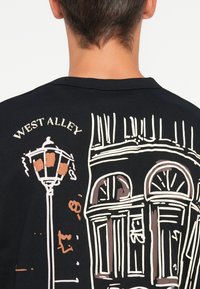 T-shirt nera a maniche corte con una grafica di un lampione e un edificio, con la scritta "WEST ALLEY" in bianco e colori tenui e morbidi.