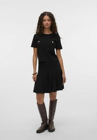 Robe en tricot noire à manches courtes, avec deux poches poitrine ornées de boutons dorés. Conception de jupe évasée, assortie à des bottes marron arrivant au genou.