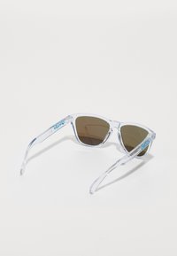 Przezroczyste plastikowe okulary przeciwsłoneczne z brązowymi soczewkami, o geometrycznym kształcie i niebieskimi akcentami logo na zausznikach. Lekka konstrukcja, odpowiednia do codziennego noszenia.