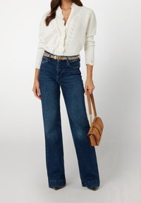 Weißes Strickbluse mit gewelltem Ausschnitt, kombiniert mit dunkelblauen Schlagjeans und einer braunen Umhängetasche mit dekorativer Stickerei.