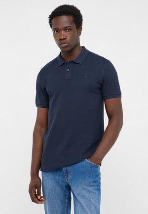 Junger Mann mit kurzen lockigen Haaren, der ein dunkelblaues Poloshirt und hellblaue Jeans trägt, steht mit einer Hand in der Tasche.