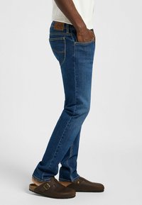 Jeans de mezclilla azul oscuro con un diseño de pierna recta, que presentan un parche de cuero marrón en la parte posterior y costuras en contraste. Llevados con zuecos marrones.