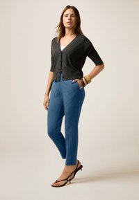 Donkergrijze cardigan met korte mouwen en knopen, gecombineerd met slim fit blauwe broeken. Geaccentueerd met gouden en kleurrijke armbanden, en zwarte sandalen.