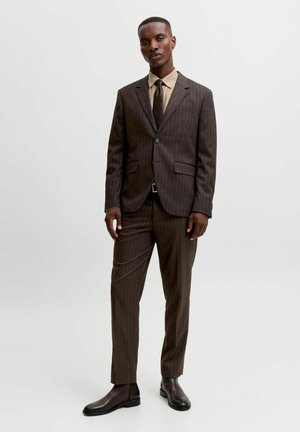 Homme debout portant un costume marron à fines rayures, une chemise beige, une cravate noire et des chaussures en cuir marron foncé sur un fond uni.