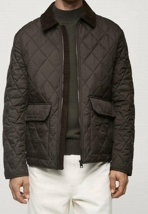 Mężczyzna ubrany w ciemnobrązową pikowaną kurtkę na oliwkowozielonym swetrze i białych spodniach, stojący z ramionami swobodnie opuszczonymi wzdłuż ciała.