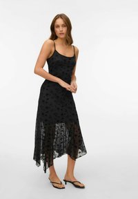 Černé šaty s květinovou krajkou, tenkými ramínky a asymetrickým lemem. Průhledná spodní vrstva dodává texturu a hloubku.