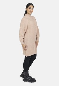 Dziergany oversize sweter w jasnoróżowym kolorze z wysokim dekoltem i prążkowaną fakturą, zestawiony z czarnymi legginsami i masywnymi czarnymi butami.