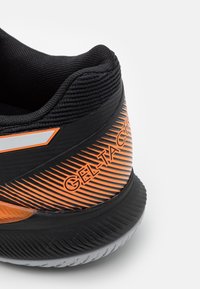 Gros plan sur le talon d'une chaussure de course noire et orange avec un col en tissu texturé et la marque "GEL-KAYANO" sur le côté.