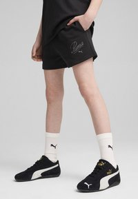 Pantalones cortos de algodón negros con el logo de Puma, combinados con zapatillas de ante negras que tienen una franja blanca y detalles en dorado, y calcetines de canalé blancos.