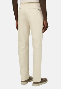 Homme portant un pantalon droit couleur crème et des chaussures plates en daim marron, vu de dos sur un fond uni.