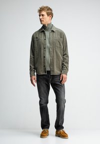 Kordjacke in gedecktem Grün, knöpfbar, mit zwei Brusttaschen, über einem mit Reißverschluss versehenen Oberteil getragen. Kombiniert mit dunklen Jeans und braunen Slippers.