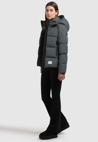 Chaqueta acolchada en gris oscuro con capucha, con cierre de cremallera y puños acanalados. Combinada con pantalones de campana negros y zapatos de plataforma.