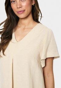 Beiges Leinenkleid mit V-Ausschnitt, kurzen rüschierten Ärmeln und Faltendetail. Glatte Textur, leichter Stoff liegt sanft an.