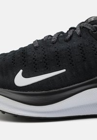 Baskets de sport noires avec tige en tissu texturé, présentant un logo swoosh blanc et des lacets gris. Design profil bas avec semelle noire.