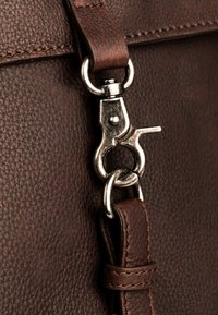 Detalle de bolsa de cuero marrón con un cierre metálico plateado, costuras resistentes y una superficie texturizada. La correa está firmemente sujeta.