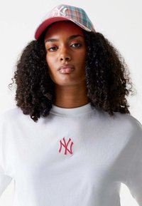 Witte T-shirt met een rood geborduurd NY-logo, gecombineerd met een meerkleurige geruiten pet met een roze klep en witte opdruk.