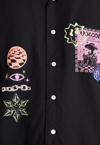 Chemise noire à boutons présentant des graphismes imprimés colorés, incluant des étoiles, des chaînes et des formes géométriques dans des tons de rose et de vert.