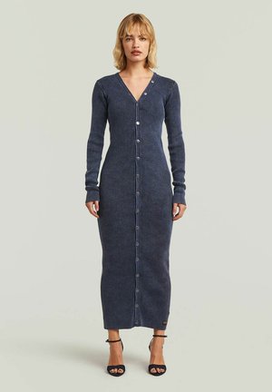 Femme portant une longue robe boutonnée côtelée bleu marine à manches longues et des talons noirs à bride à la cheville, debout devant un fond clair uni.