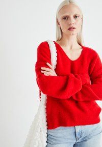 Pull en tricot rouge et duveteux avec un large encolure et des manches longues. Associé à un jean en denim bleu clair et un sac blanc texturé.