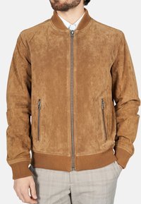 Man in een lichtbruine suede bomberjack met geribde manchetten en zoom, ritssluiting aan de voorkant, en een lichtgrijze geblokte broek, met de handen gedeeltelijk in de zakken.