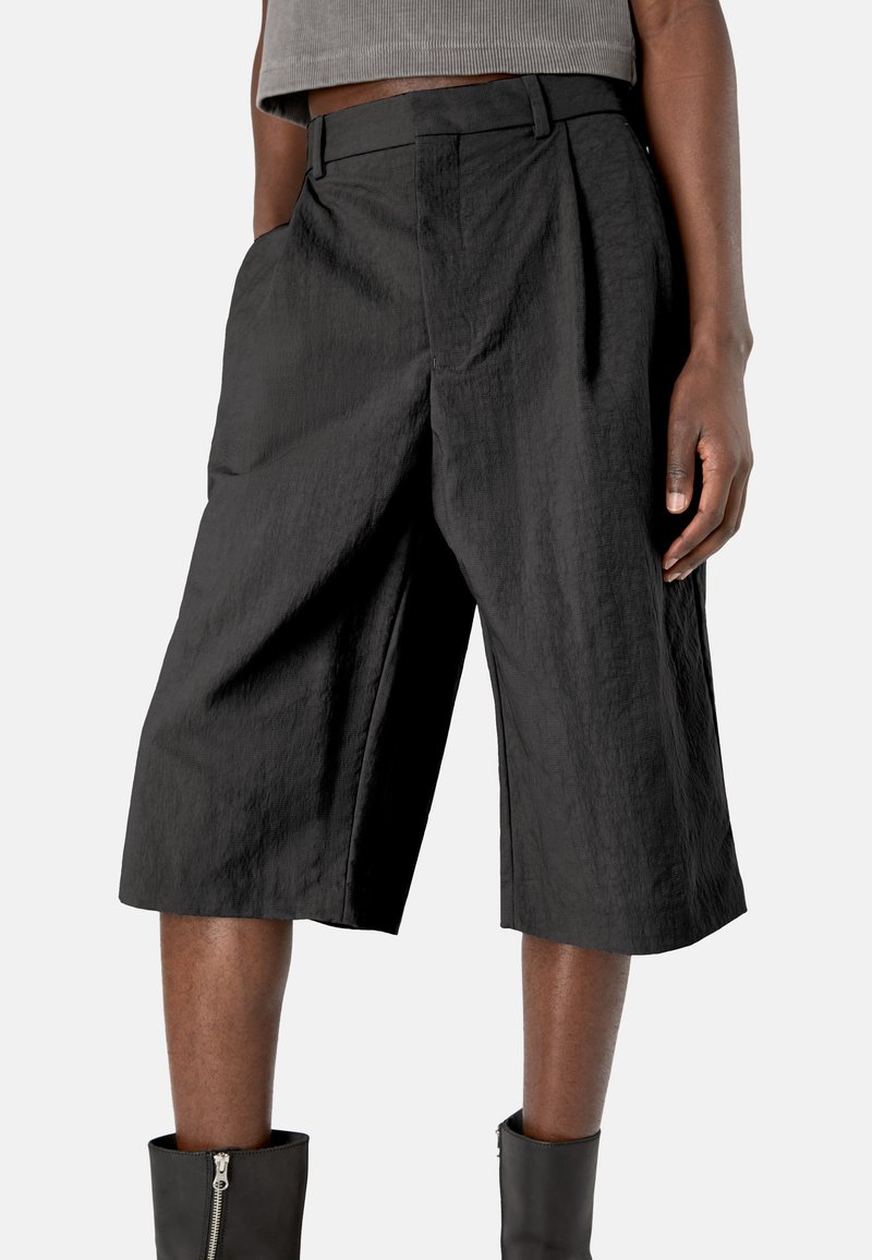 Pantalones cortos negros de longitud media hechos de una tela texturizada con un diseño plisado. Cuenta con bolsillos laterales y una cintura ajustada, combinados con botas de tobillo.