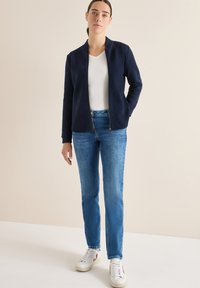 Námořnická bunda na zip s texturovaným povrchem, doplněná světle modrými džíny s rovnými nohavicemi a bílými teniskami s barevnými akcenty.