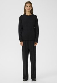 Eine Frau steht in einem schwarzen Strickpullover, einer schwarzen weit geschnittenen Hose und schwarzen Schuhen vor einem weißen Hintergrund.