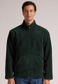 Zielona polarowa kurtka na zamek z teksturowaną powierzchnią, stojącym kołnierzem i kieszeniami z przodu. Luźny krój z długimi rękawami.