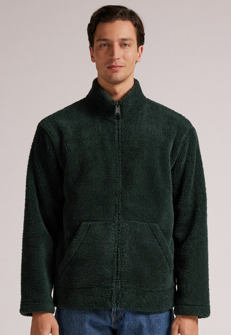 Zielona polarowa kurtka na zamek z teksturowaną powierzchnią, stojącym kołnierzem i kieszeniami z przodu. Luźny krój z długimi rękawami.